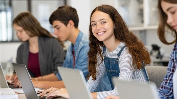 „2050 – Es liegt bei dir“: Europas Zukunft spielend gestalten Eine Schülerin lächelt während sie an ihrem Laptop arbeitet. Um sie herum sitzen weitere Schüler*innen, die ebenfalls an deren Laptop arbeiten.