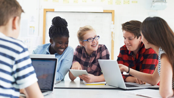 Mehrer Schüler sitzen vor Laptops/Tablets und unterhalten sich.