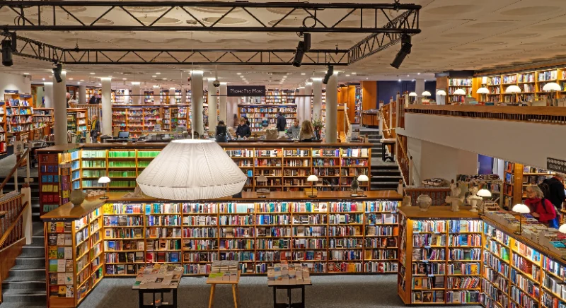 Auf dem Bild ist ein großzügiger Bücherladen mit zwei Etagen zu sehen. Die Regale haben eine Holzoptik und in der Mitte des Raumes hängt eine etwa 2 Meter breite Lampe.