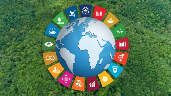 Die Erde erhebt sich über einen grünen Wald. Um sie herum sind Icons zu sehen, die verschiedene Nachhaltigkeitsziele illustrieren: Familienleben, Geschlechtergerechtigkeit, nachhaltige Energieproduktion, Frieden und so weiter